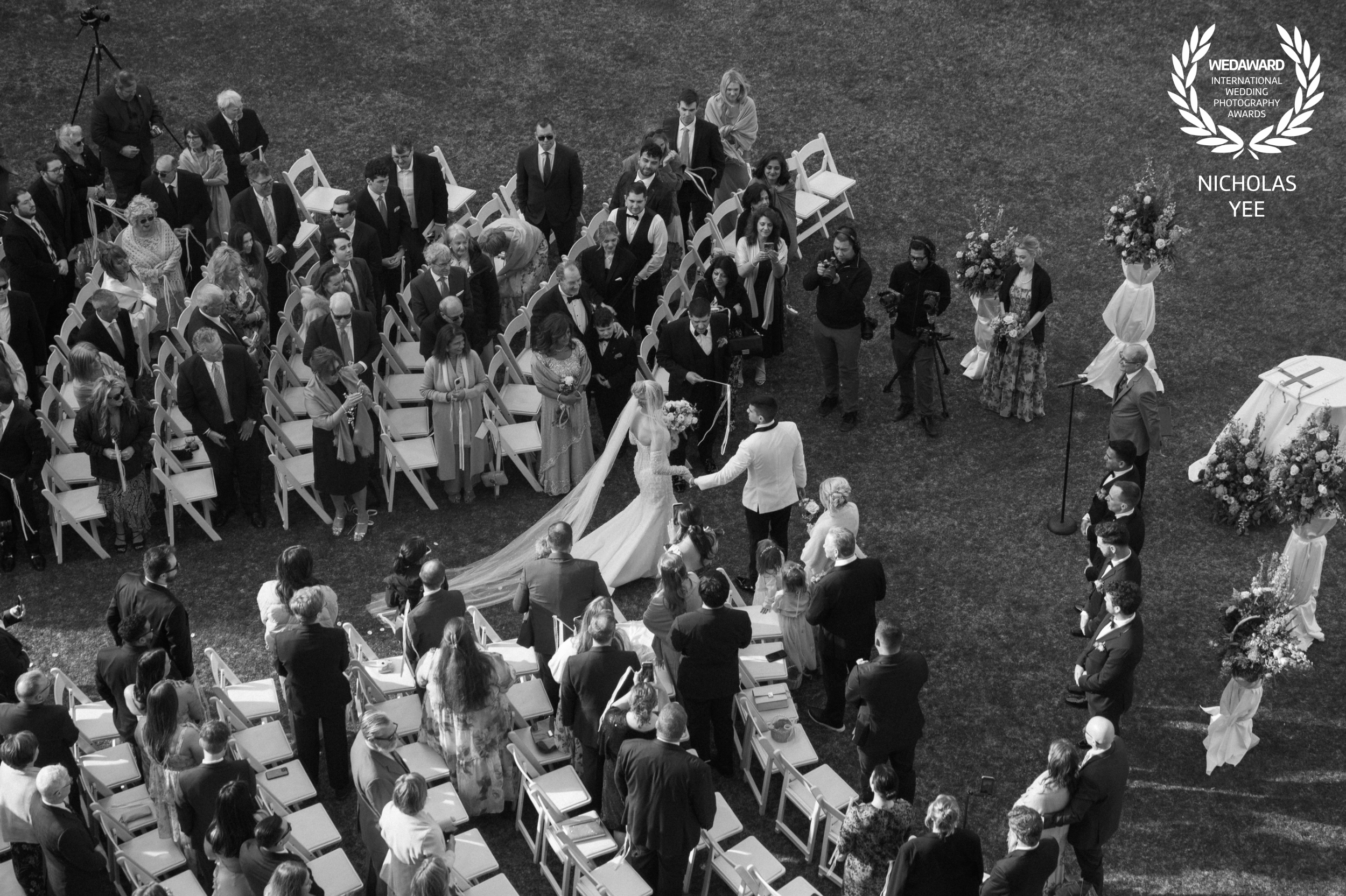 Overhead black-and-white photograph of newlyweds holding hands in an outdoor ceremony aisle surrounded by standing guests.