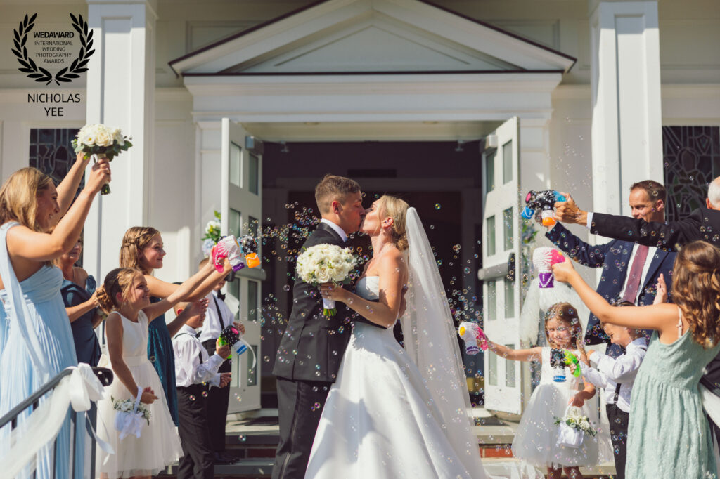 Newlyweds kissing outside a white church while family members and young wedding attendants surround them with bubbles.