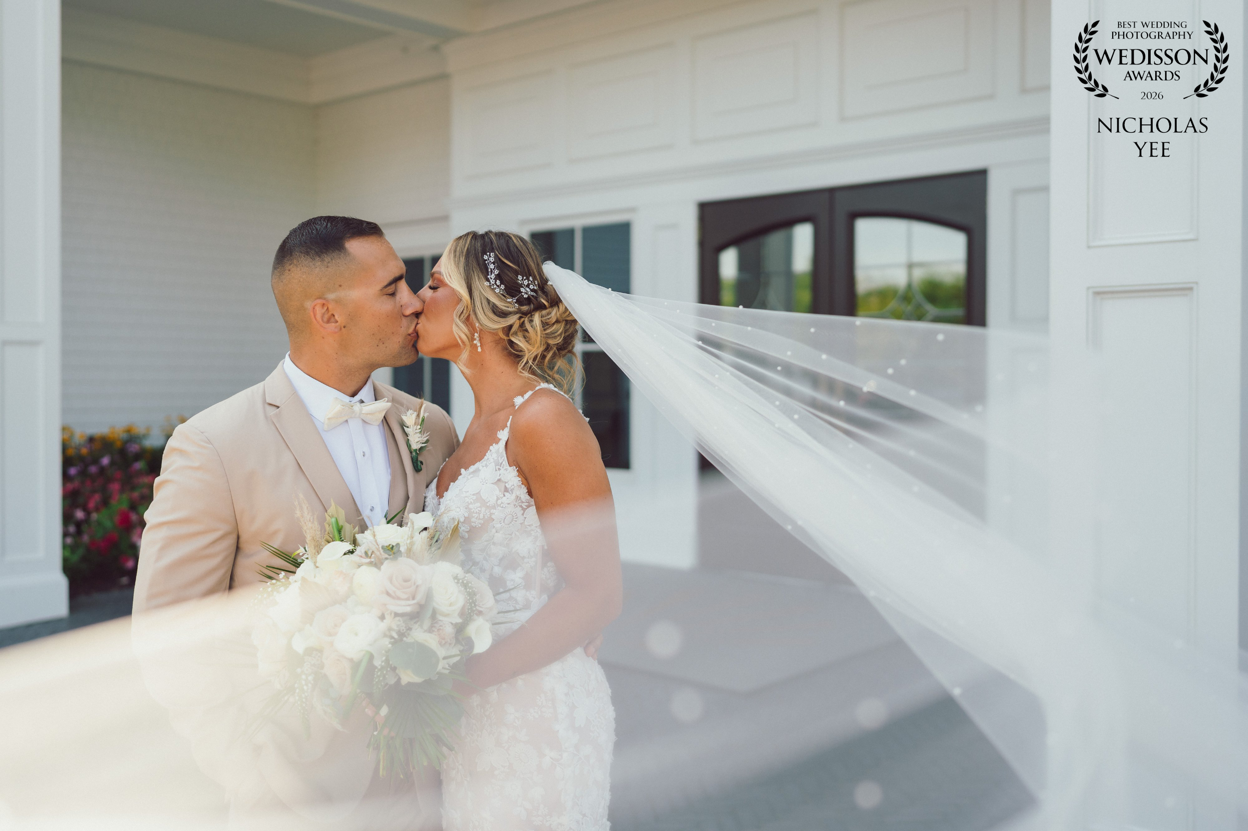 Bride and groom kissing outside a white wedding venue as the bride’s long veil sweeps across the frame, with her white floral bouquet in the foreground.