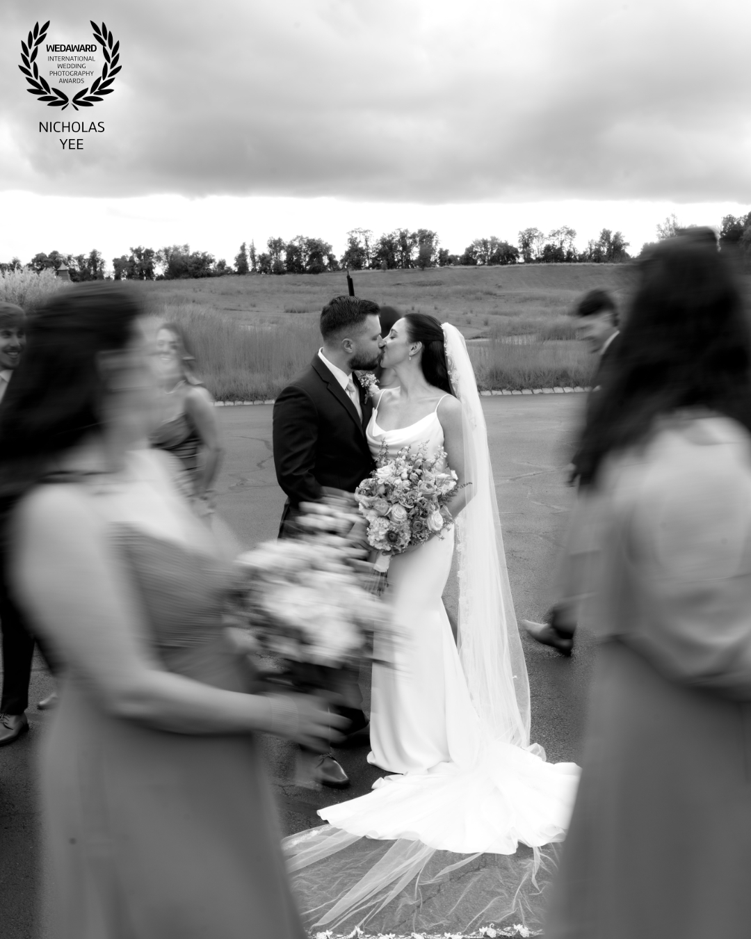 Black-and-white photo of a bride and groom kissing while their wedding party moves around them in a soft blur.